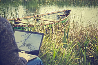 Person Working on Laptop by Lake with Wooden Boat
