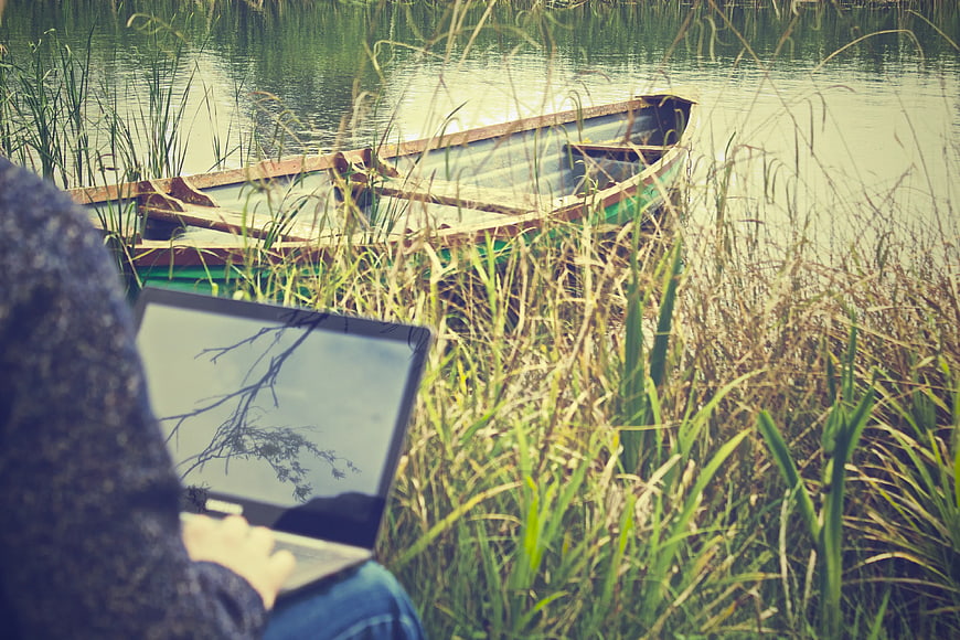 Person Working on Laptop by Lake with Wooden Boat