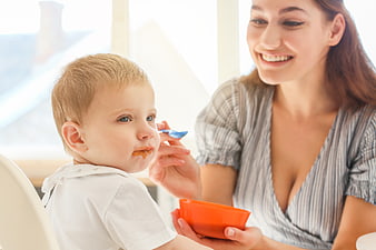 Happy mother feeding baby with orange bowl indoors