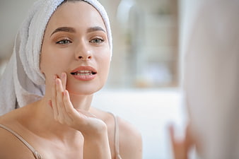 Woman with towel on head touching face in bathroom mirror