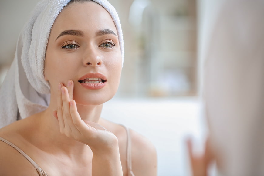 Woman with towel on head touching face in bathroom mirror