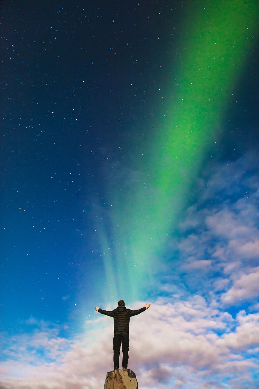 Man with arms raised under green aurora borealis night sky