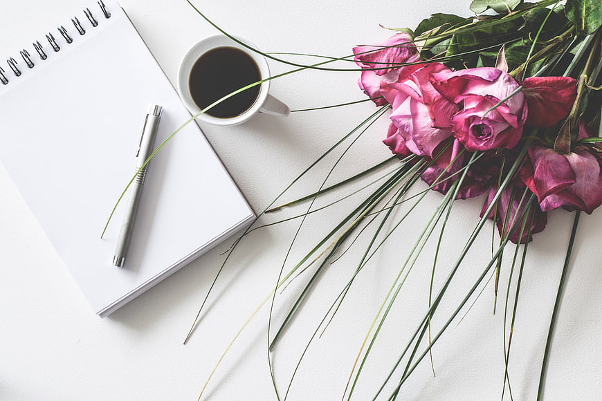 Flat lay of pink roses, coffee cup, and notepad on white background