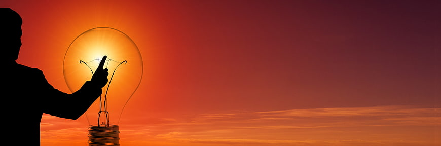 Silhouette of man touching lightbulb against sunset sky