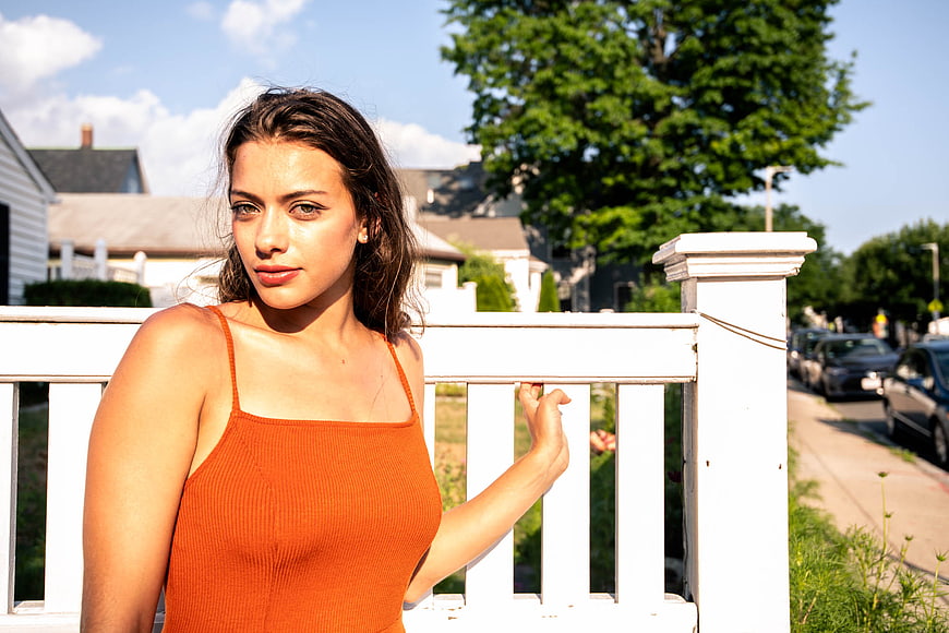 Young Woman in Orange Top Leaning on White Fence Outdoors