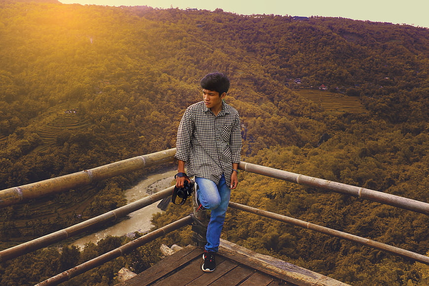 Young man with camera on bamboo bridge overlooking lush valley
