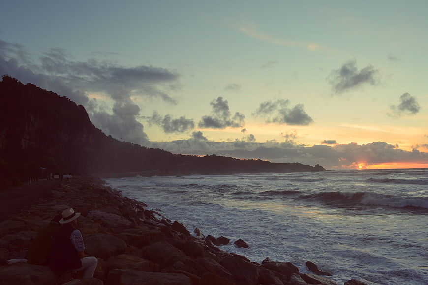 Person sitting on rocks watching sunset over ocean waves