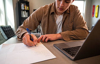 Young man writing notes on paper at desk with laptop