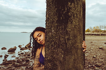 Woman with tattoos peeking from behind a tree on a rocky beach