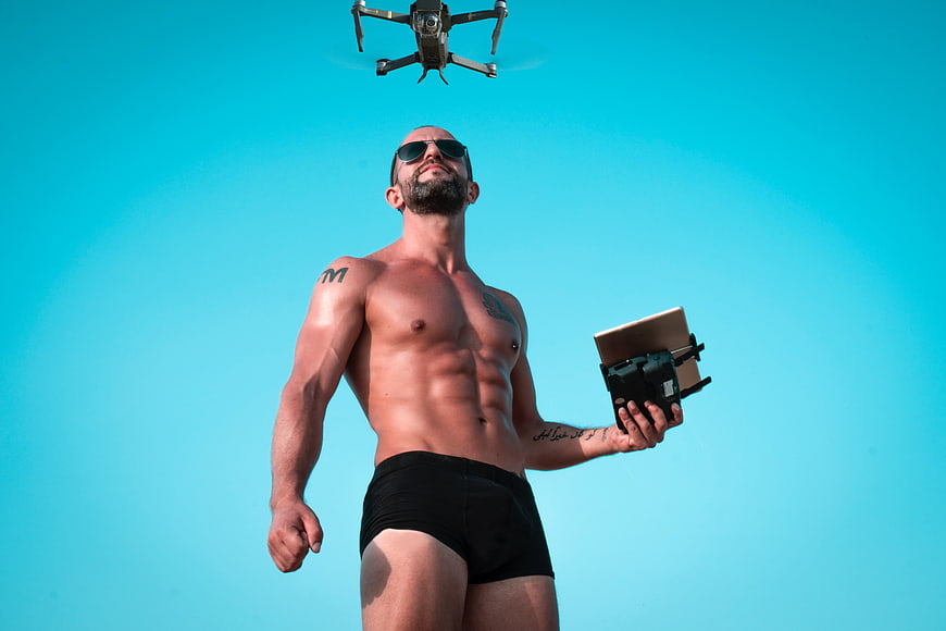 Shirtless muscular man flying drone against blue sky