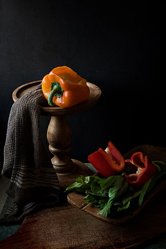 Dark moody still life of orange and red bell peppers on wooden stand