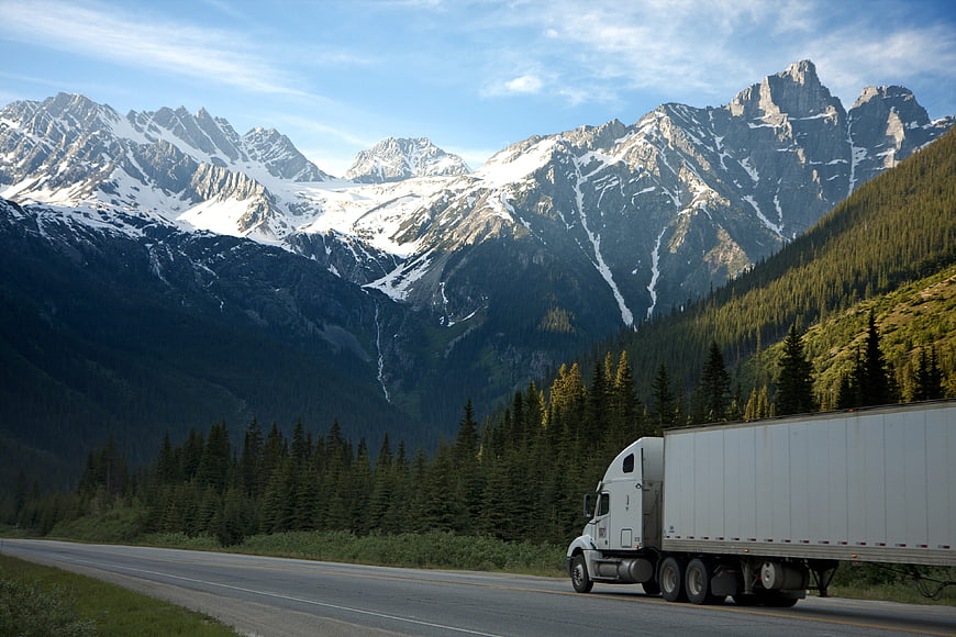 White semi-truck driving on highway with snow-capped mountains