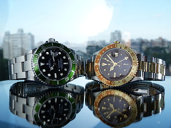 Two luxury wristwatches with green and gold bezels on a reflective surface