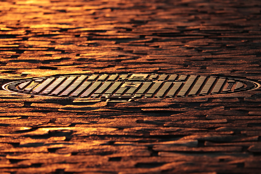 Golden Manhole Cover On Wet Cobblestone Street At Sunset