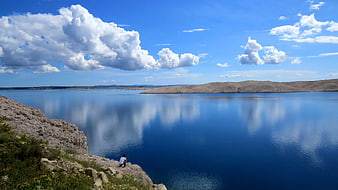 Solitary figure overlooking vast blue lake with cloud reflections