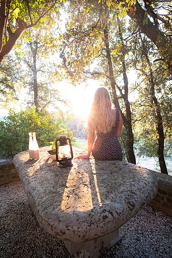 Woman sitting on stone bench in sunlit forest with lantern