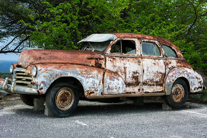 Rusty vintage car parked on pavement with green trees