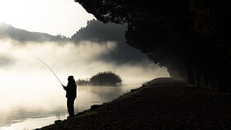 Silhouette of fisherman on misty river bank at dawn