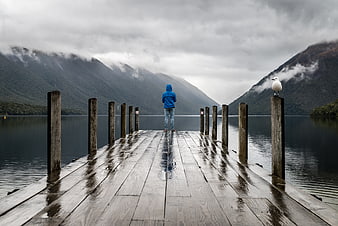 Lone figure in blue hoodie on wet wooden pier with seagull