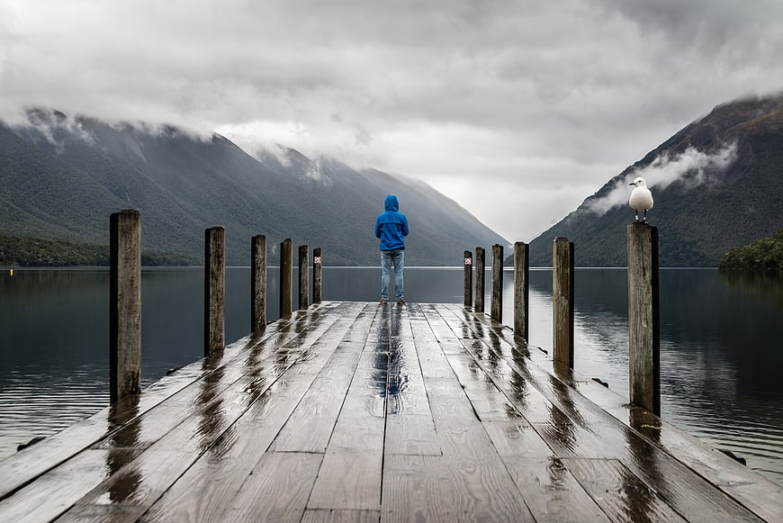 Lone figure in blue hoodie on wet wooden pier with seagull