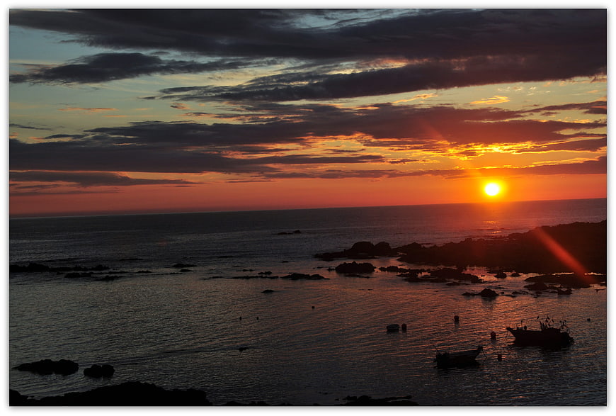 Dramatic sunset over rocky coastline with silhouetted boats
