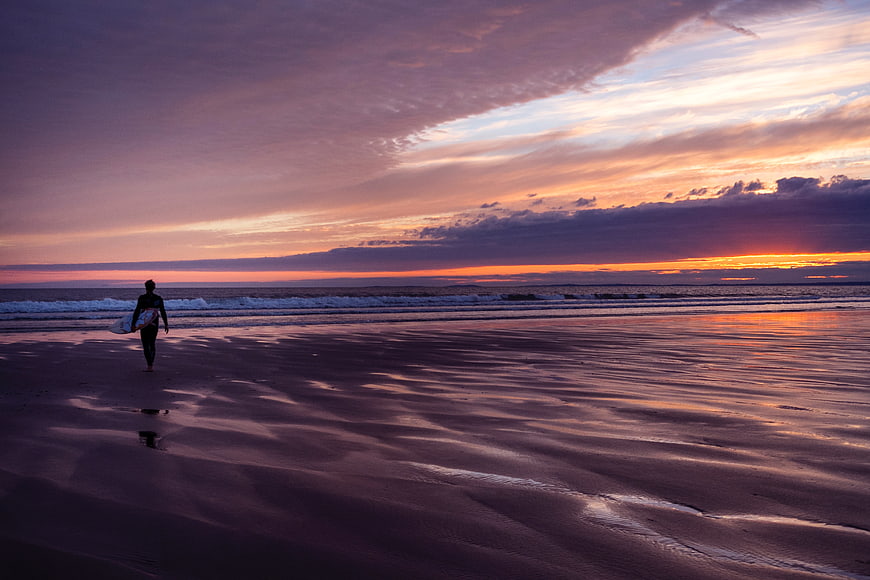 Surfer Walking on Beach at Sunset with Purple Sky