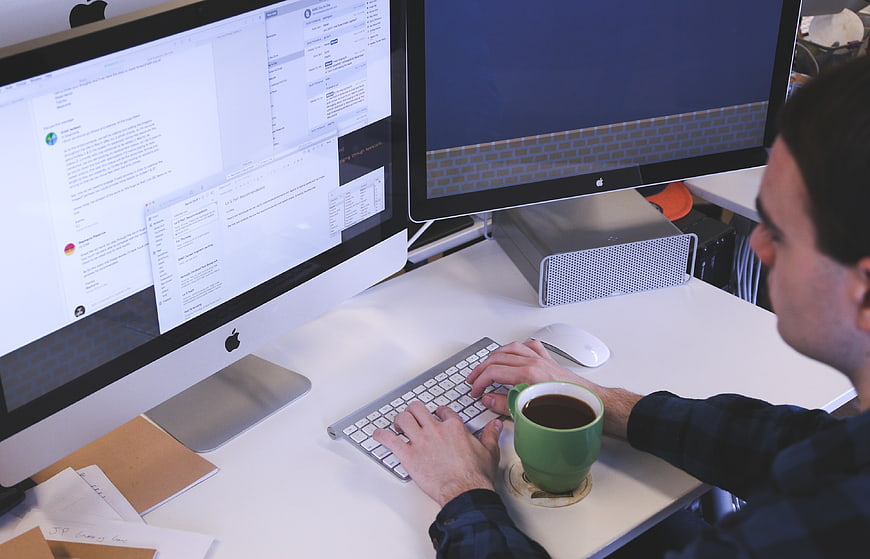 Man Typing at Dual Monitor Workspace with Coffee