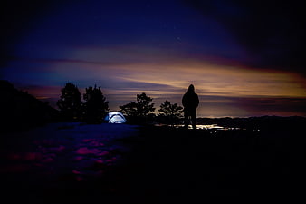 Silhouette of camper with glowing tent overlooking city lights at night