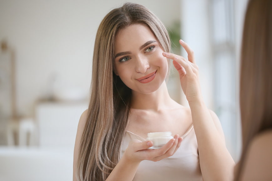 Young woman applying face cream in bathroom mirror