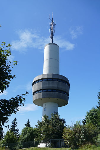 Modern white broadcast tower against blue sky with trees