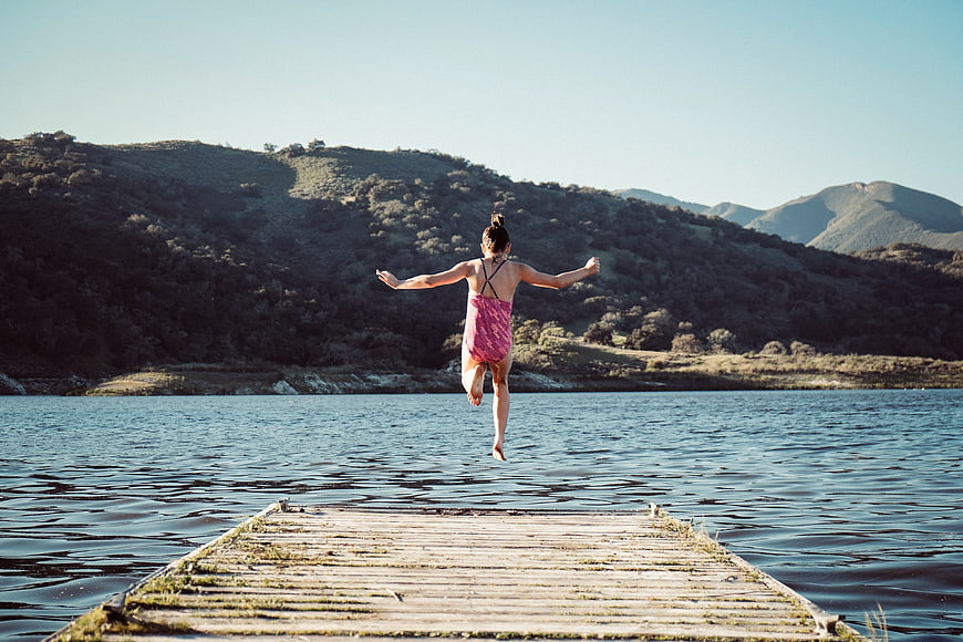 Woman jumping off wooden dock into lake with mountains