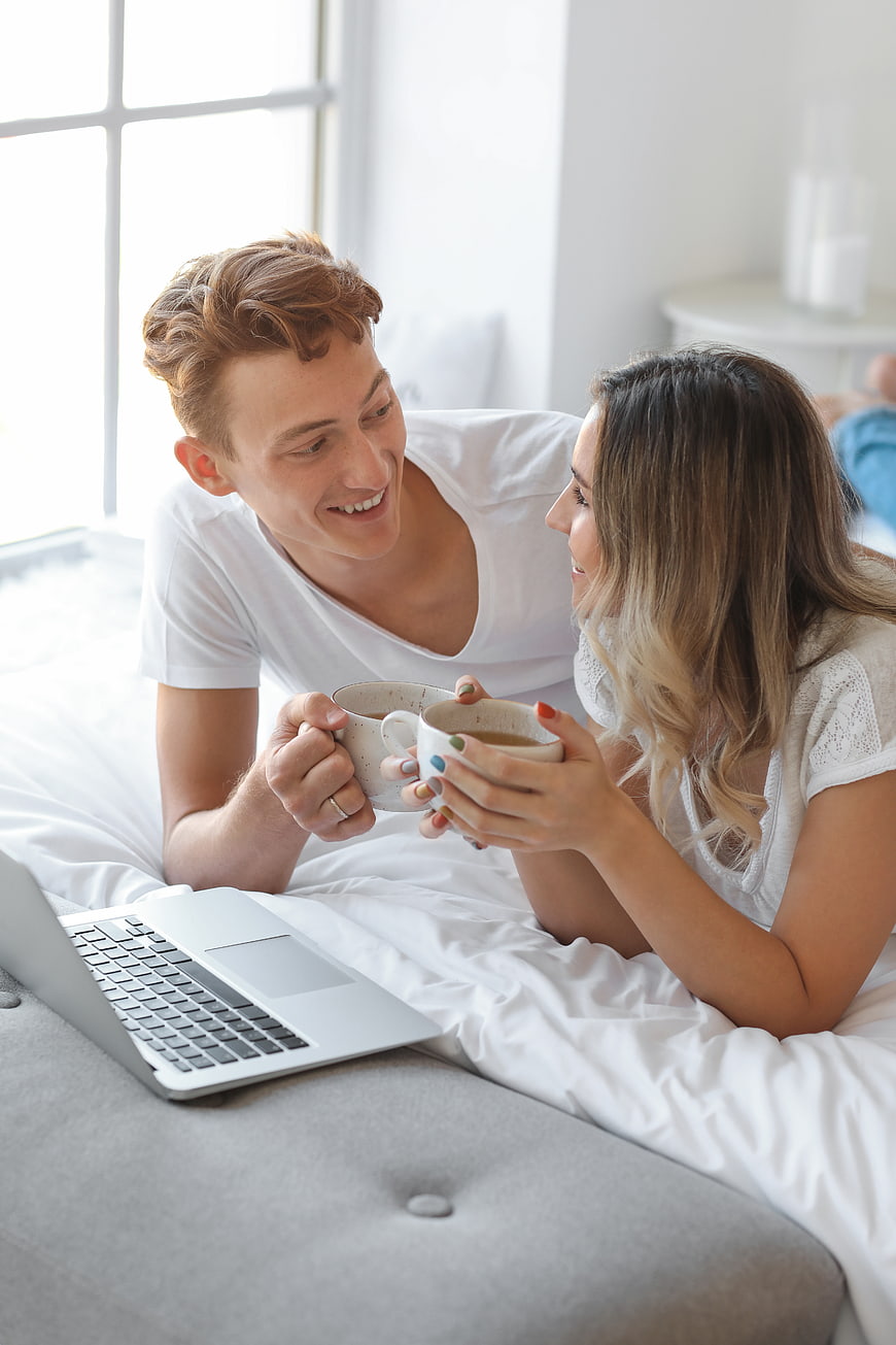 Happy young couple drinking coffee in bed with laptop