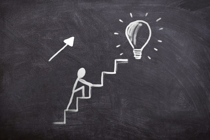 Chalk drawing of person climbing stairs toward lightbulb on blackboard