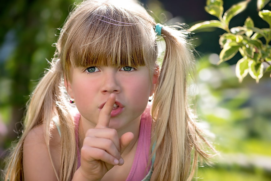 Cute blonde girl with pigtails shushing finger to lips outdoors