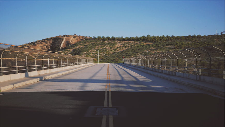 Empty concrete bridge road with chain-link fence and hills
