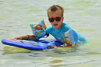 Young boy with sunglasses and floaties on a blue bodyboard in the ocean
