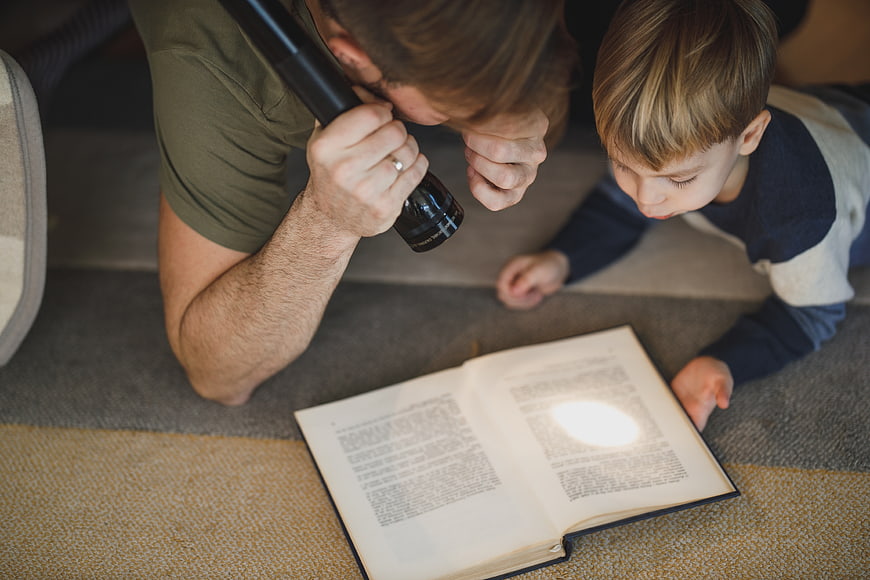 Father and son reading book with flashlight on carpet