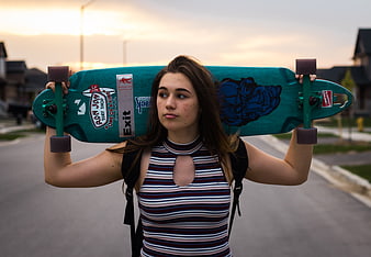 Teenage girl holding teal longboard on suburban street at sunset