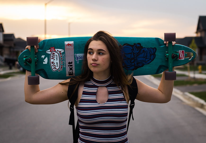 Teenage girl holding teal longboard on suburban street at sunset