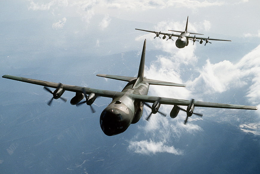 Two vintage military propeller planes flying in formation over a mountainous landscape