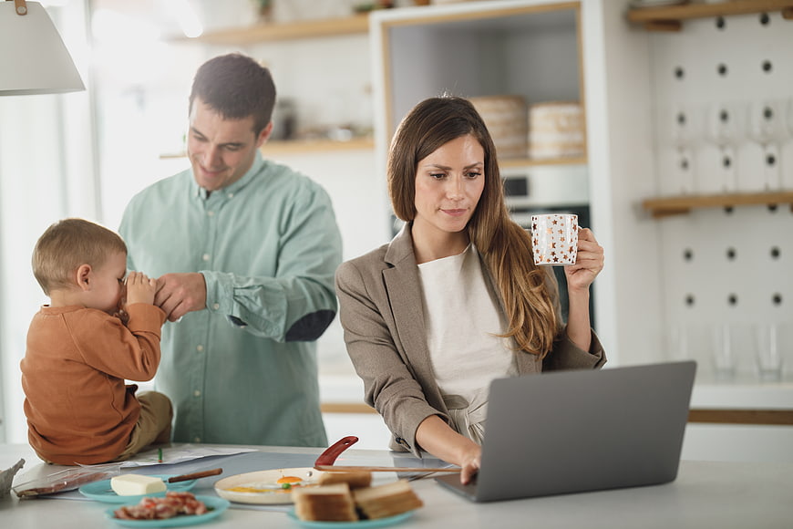 Mother working on laptop while father plays with toddler at breakfast