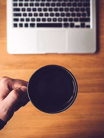 Hand holding black coffee cup over laptop on wooden desk