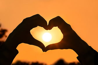 Silhouette of hands forming a heart shape against a bright orange sunset sky