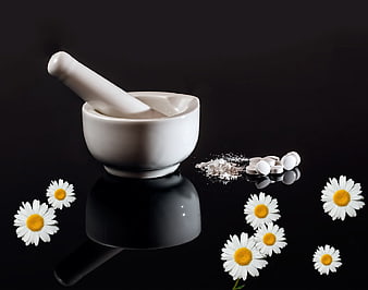 White mortar and pestle with chamomile flowers and pills on black background