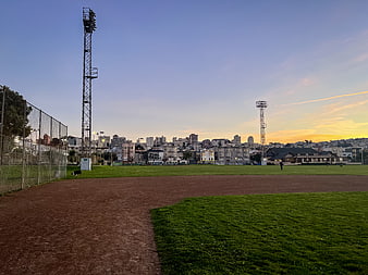 Urban baseball field at sunset with city skyline background