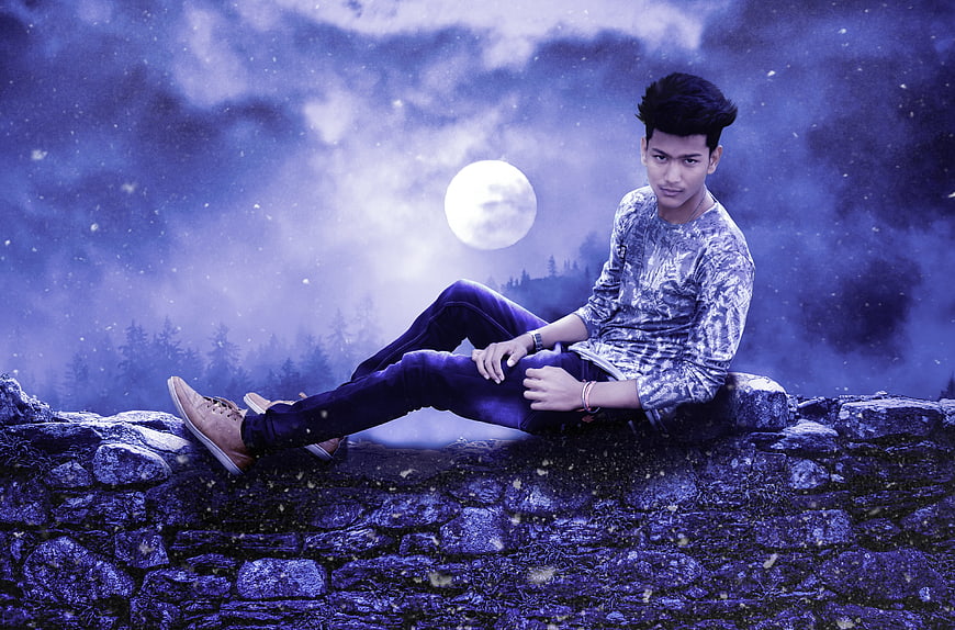 Young man sitting on stone wall under full moon night sky