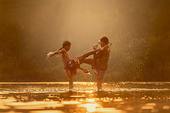 Two boys practicing Muay Thai kickboxing in water at sunset