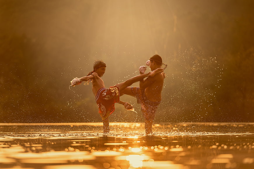 Two boys practicing Muay Thai kickboxing in water at sunset