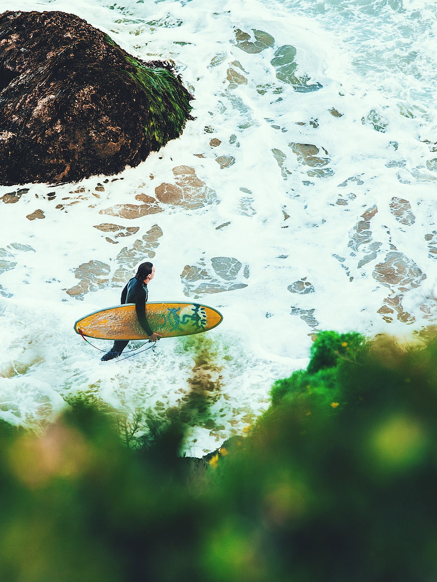 Surfer with yellow board walking through ocean waves near rocks