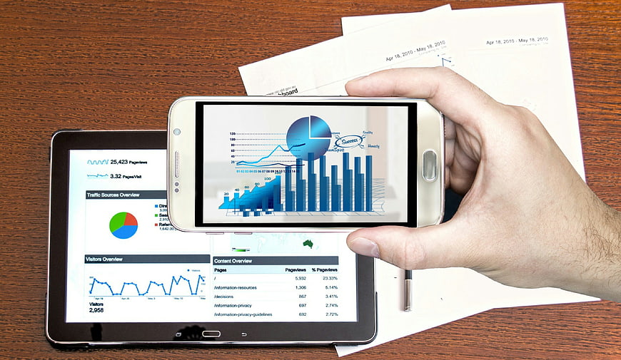 Hand holding smartphone with analytics charts over tablet and printed reports